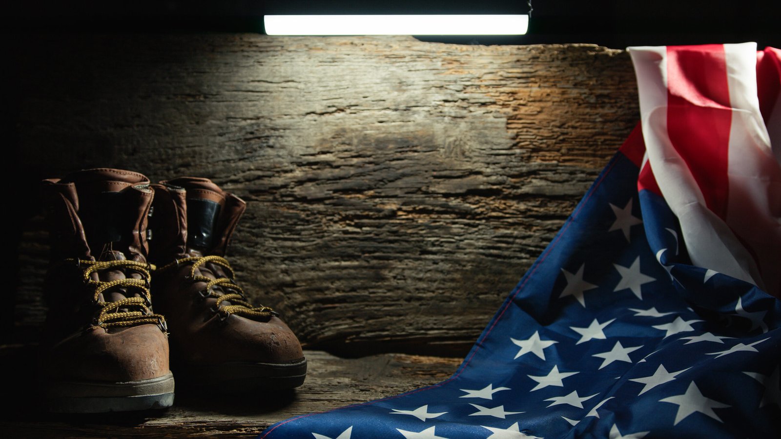 Ein Paar abgenutzte braune Kampfstiefel stehen auf einer Holzfläche unter einem hellen Licht. Rechts hängt eine gefaltete amerikanische Flagge über dem Holz, die vom Licht beleuchtet wird. Der Hintergrund ist eine schroffe, strukturierte Holzwand, die der Szene eine rustikale und feierliche Atmosphäre verleiht.
